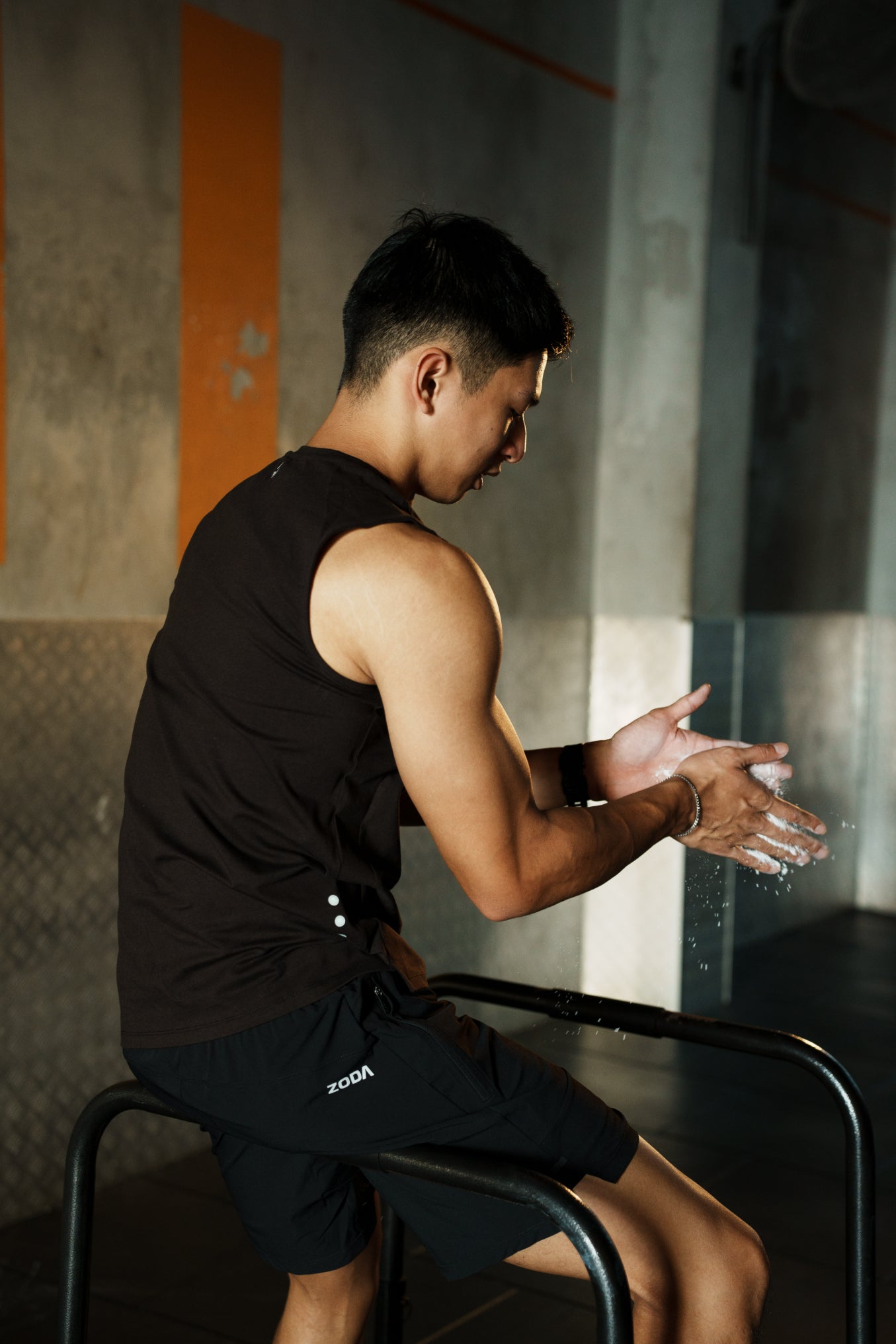 A man sitting on a bench, wearing black ZODA perform shorts and a sleeveless top, clapping his hands together. zoda.sg