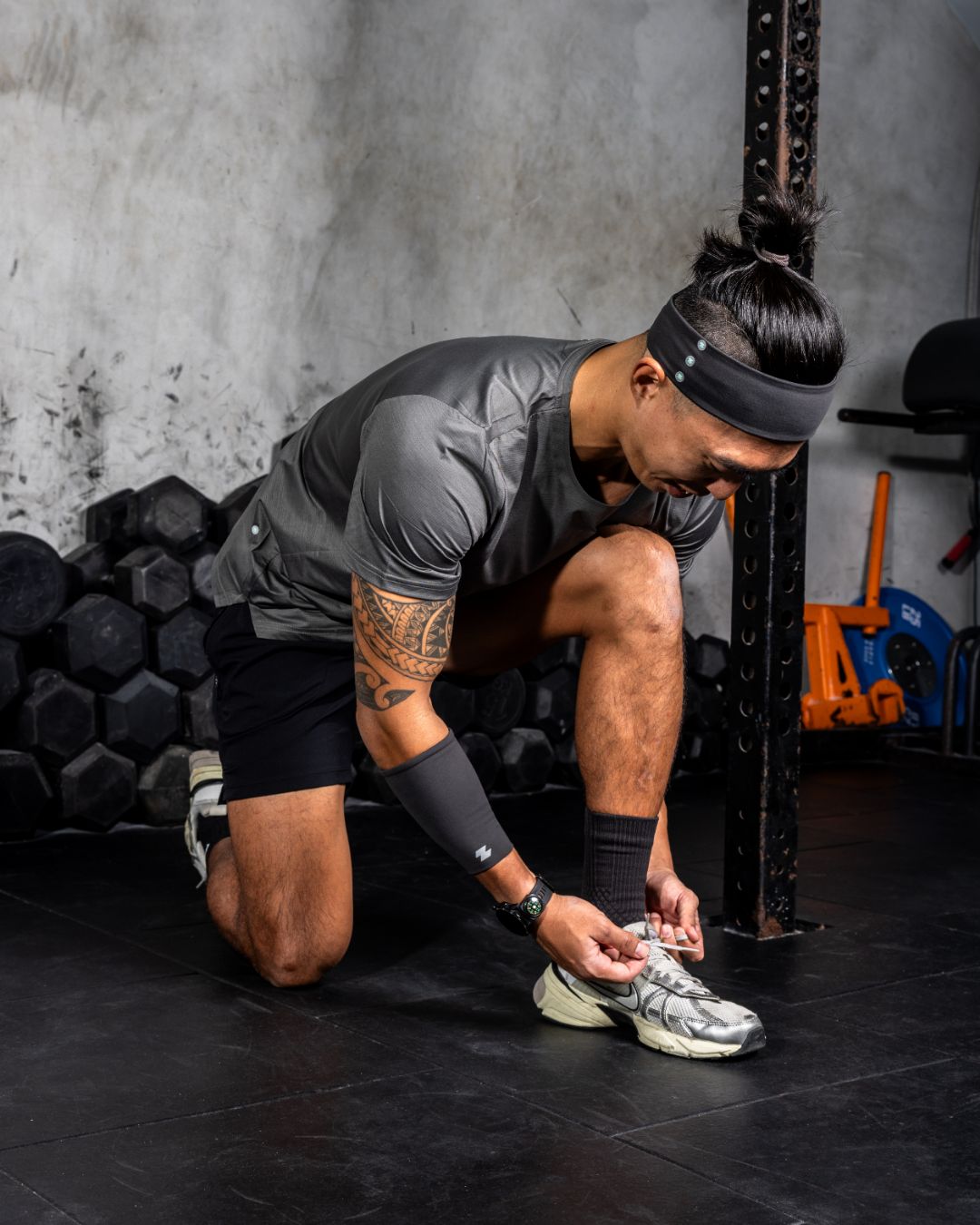 Person tying their shoe in a gym setting with weights and exercise equipment in the background. Zoda.sg