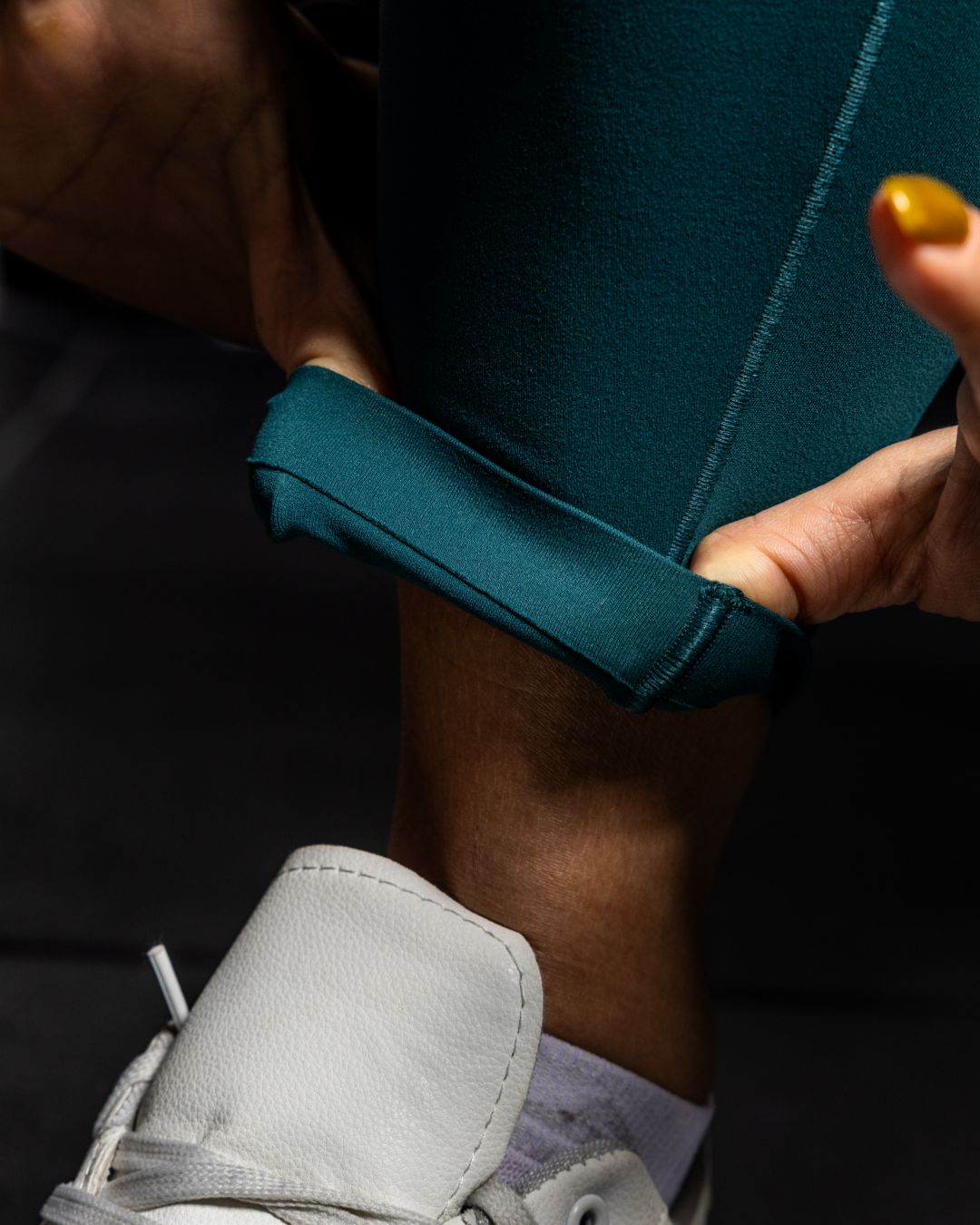Person wearing teal pants with rolled-up cuffs and gray sneakers on a dark background Zoda.sg