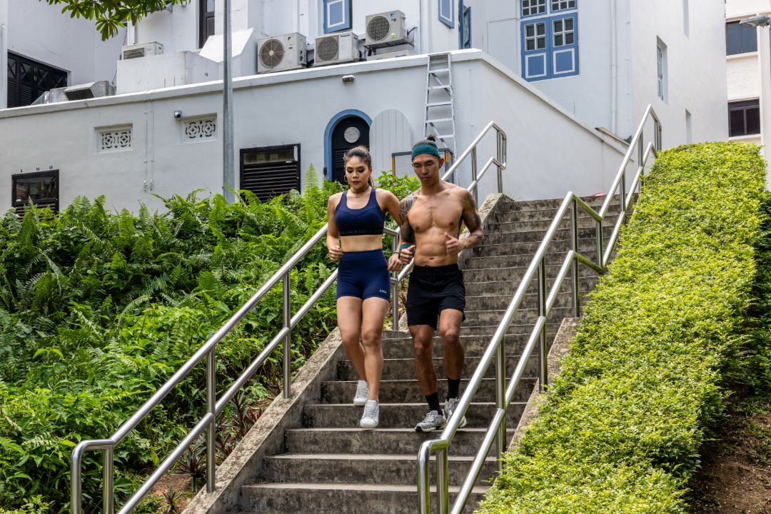 Two people in athletic wear running up a set of stairs with a white building and greenery in the background. Zoda.sg