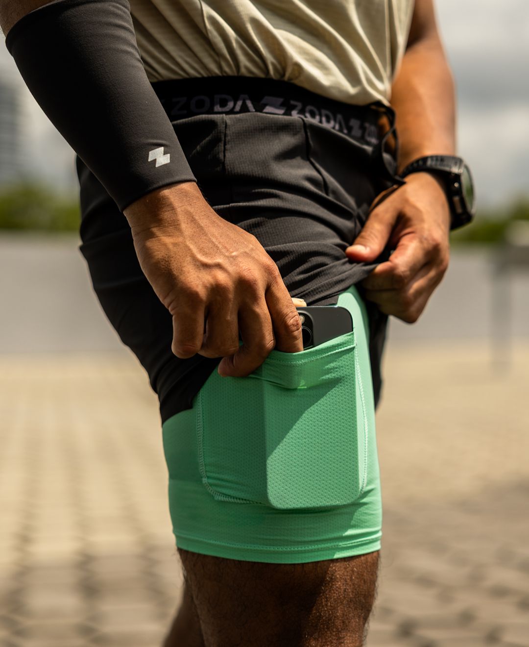 Person wearing green athletic shorts with a pocket, black arm sleeve, and beige shirt. Zoda.sg