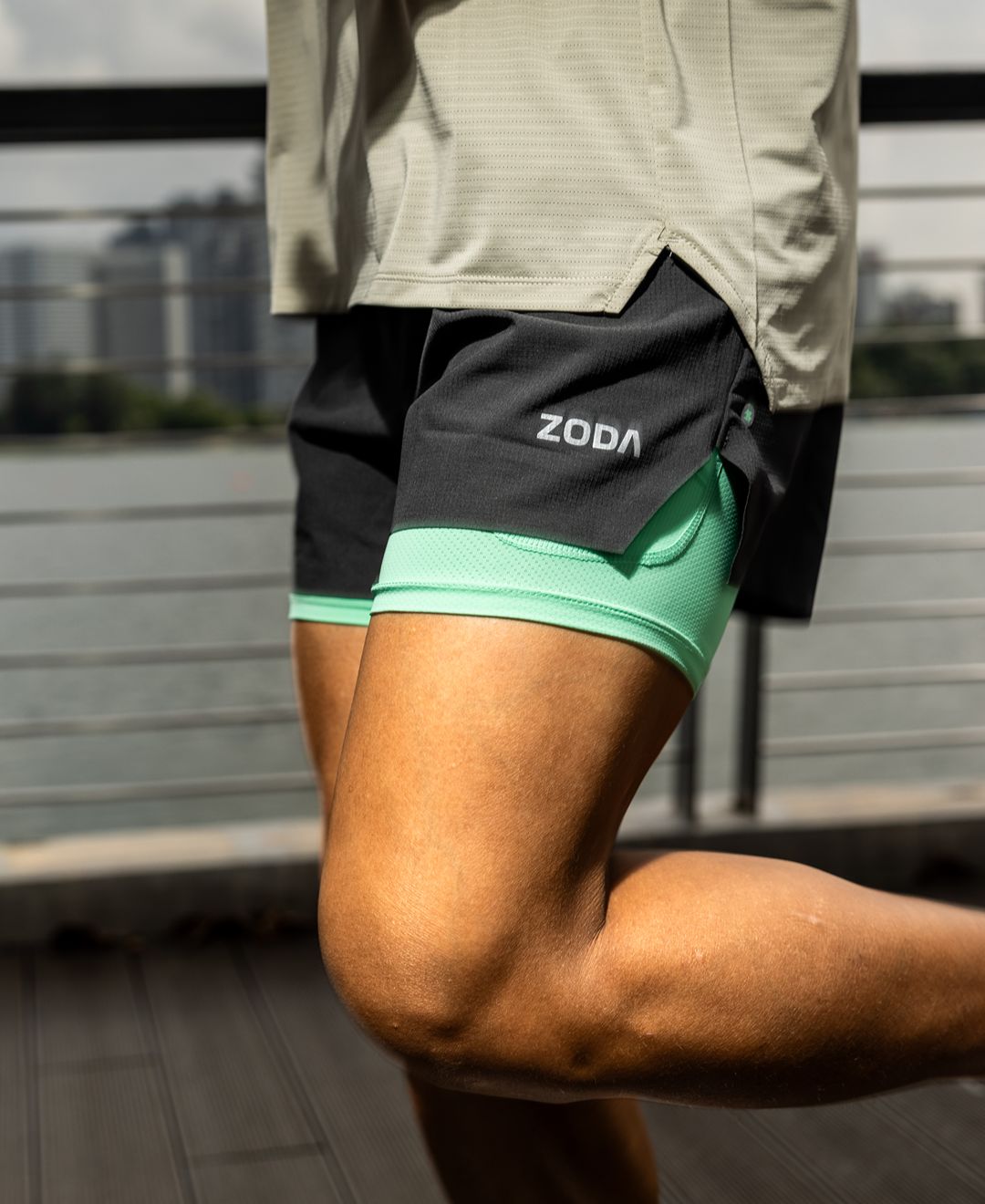 Person wearing black and green athletic shorts with 'Zoda' branding on a blurred outdoor background. Zoda.sg