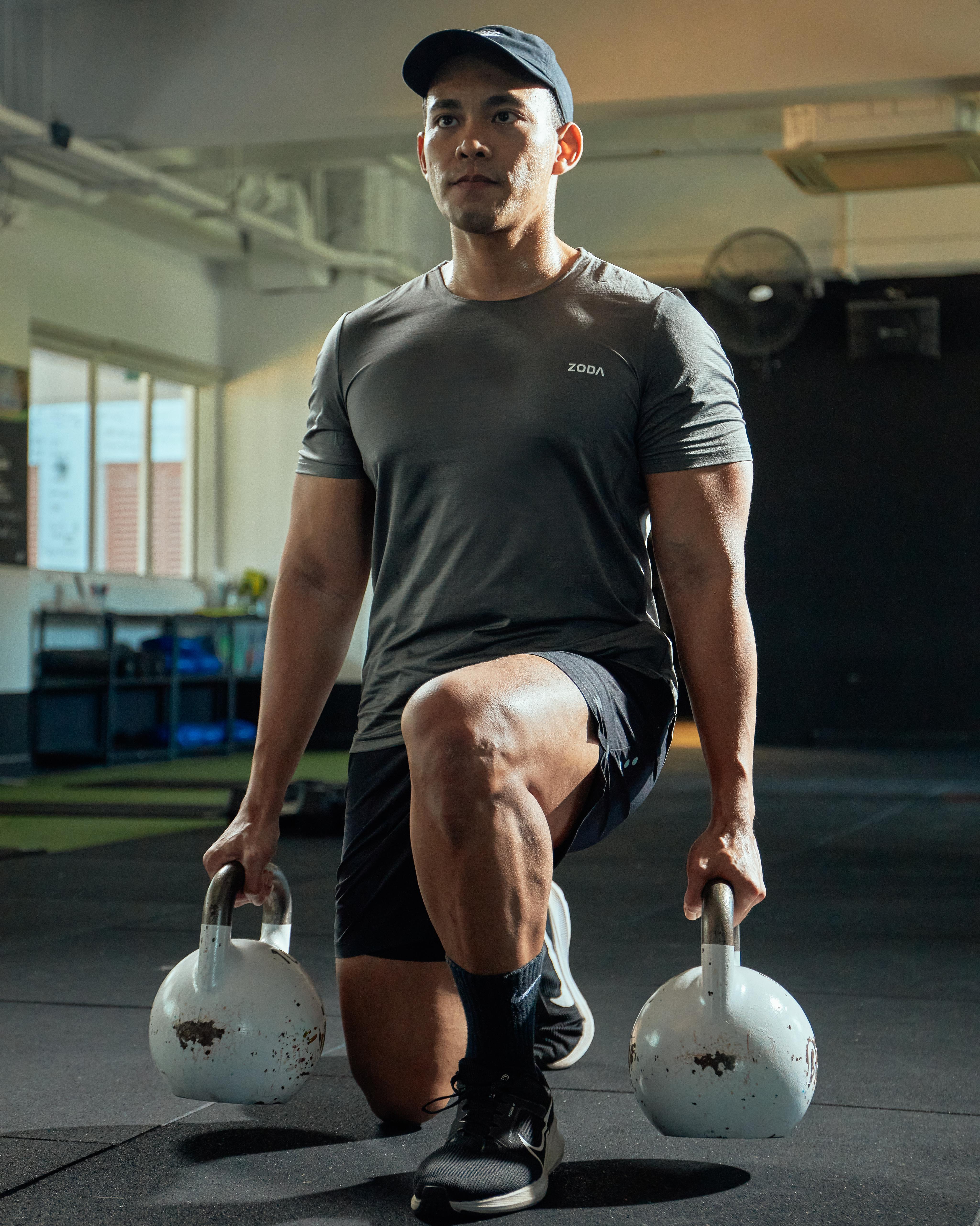 A man performing a workout holding two kettlebells, wearing a gray performance tee and black shorts. Zoda.sg