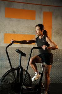 A woman engaging in exercise bike training wearing black ZODA perform shorts and a sports top, with a focus on her form and technique. Zoda.sg