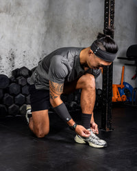 Person tying their shoe in a gym setting with weights and exercise equipment in the background. Zoda.sg