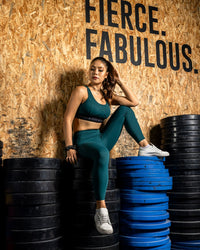 Woman in green athletic wear sitting on stacked weight plates with 'FIERCE. FABULOUS.' text on a wooden wall. Zoda.sg