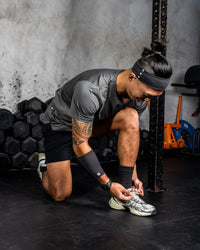 Zoda.sg Man tying his shoe in a gym setting with weights and exercise equipment in the background.